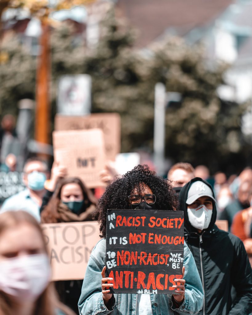 Black woman marching with protestors with a sign that reads, "in a racist society it is not enough to be non-racist, we must be anti-racist #BLM"